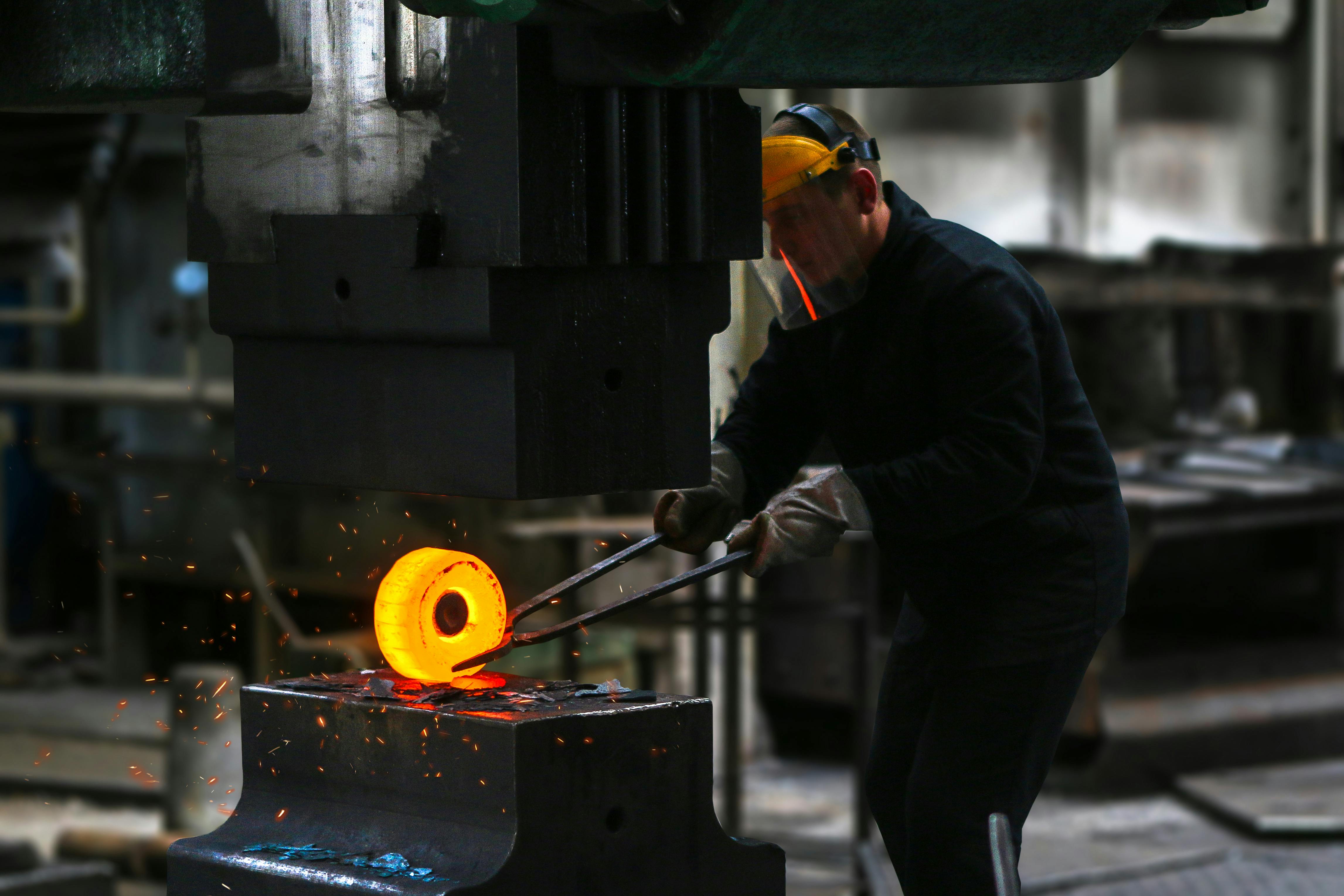 A worker uses tongs to hold a glowing, hot metal ring under a large industrial machine in a factory setting.