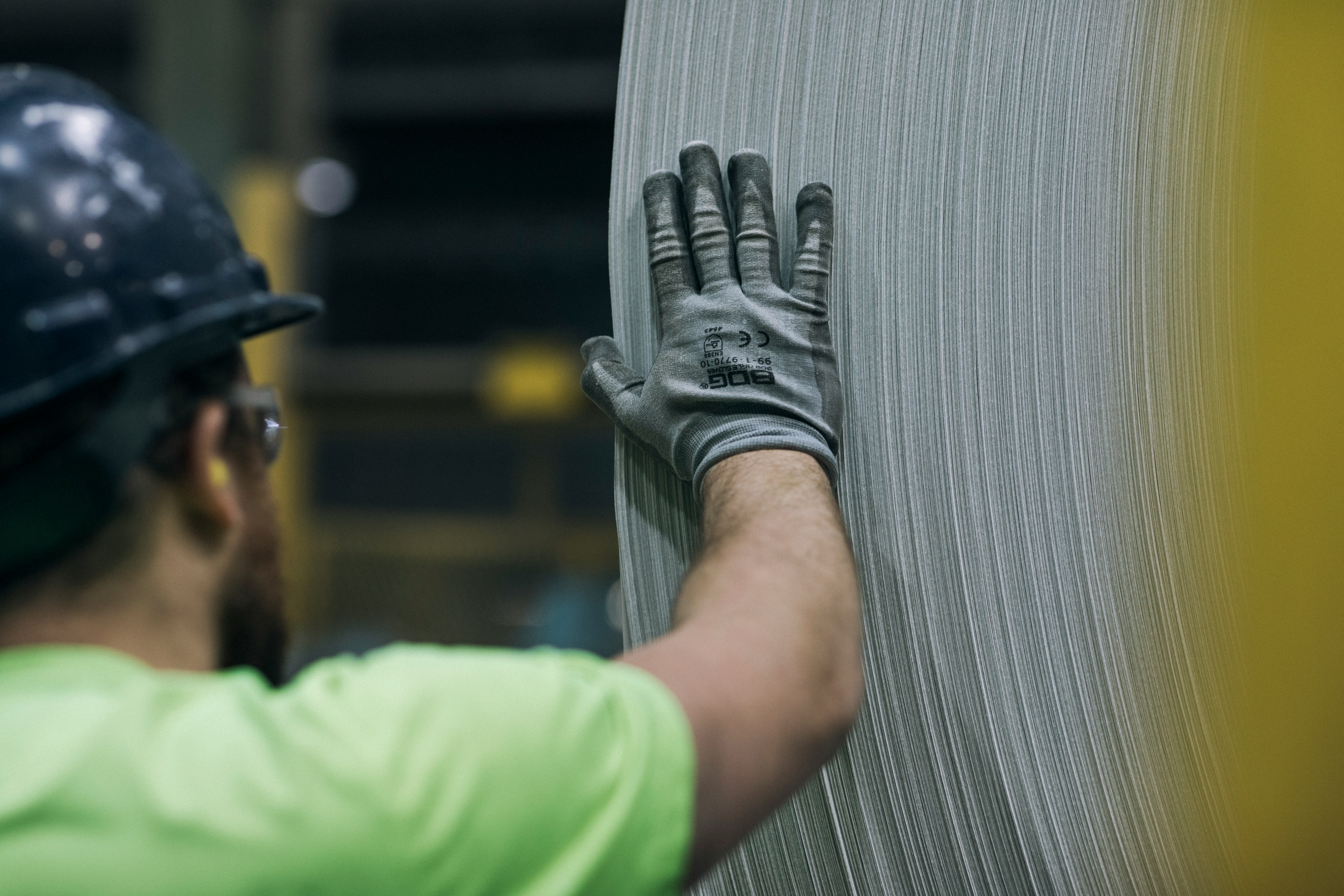 Worker in a safety helmet and gloves inspecting or guiding a large roll of sheet metal in an industrial setting.