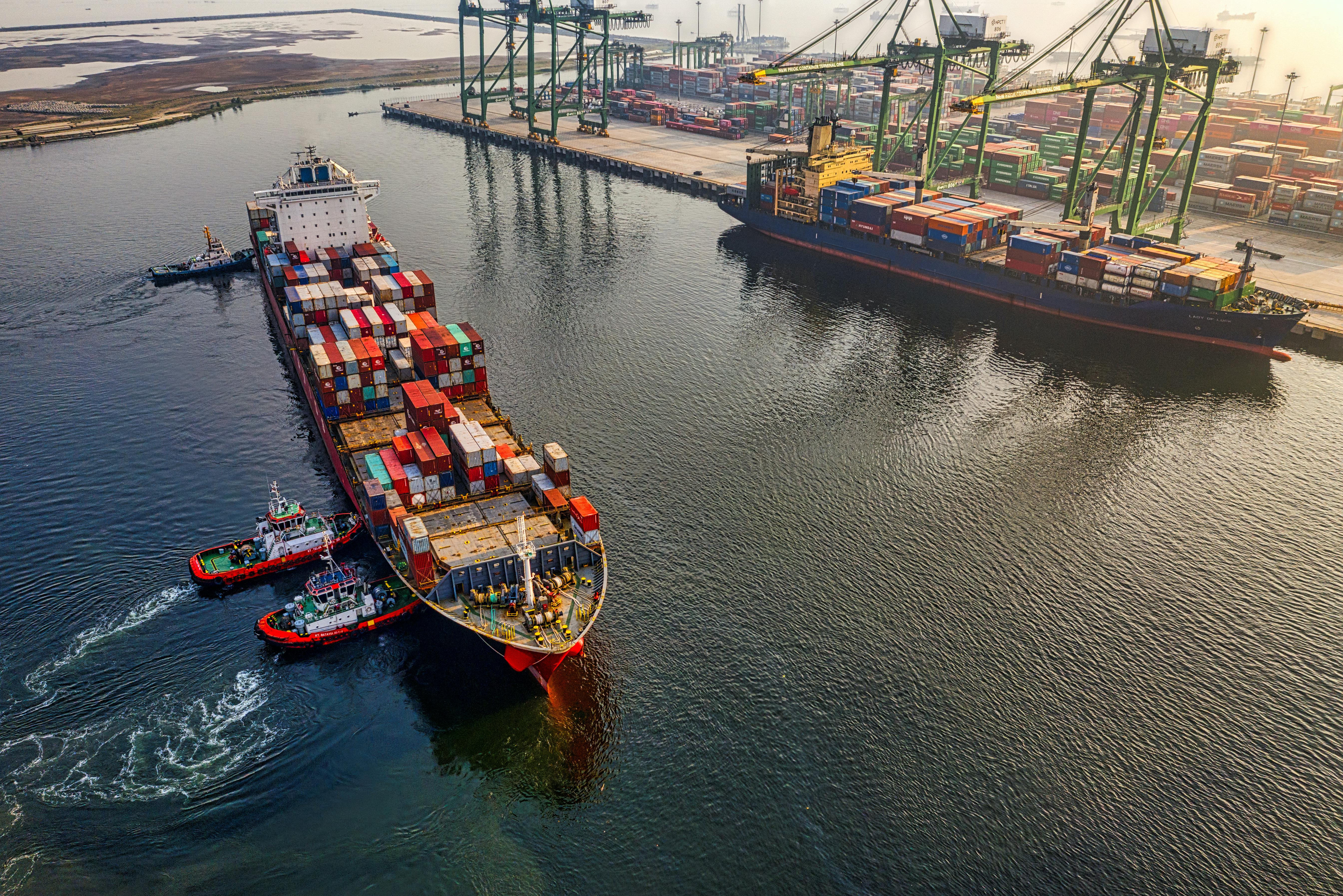 An aerial view of a busy port shows a large container ship maneuvering with assistance from two tugboats.