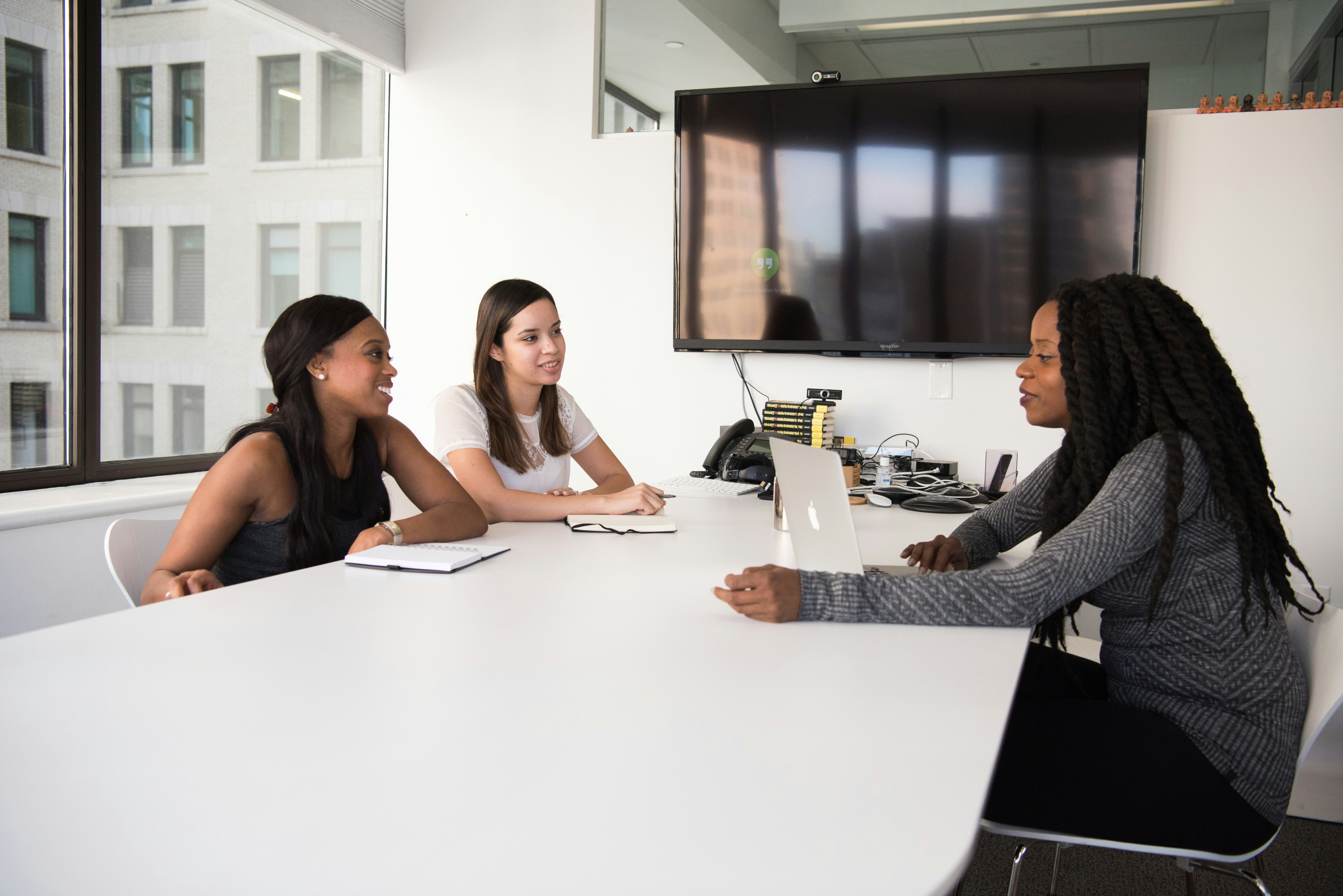 Three women sitting around a conference table in a modern office, engaged in a meeting with a laptop and notepads.