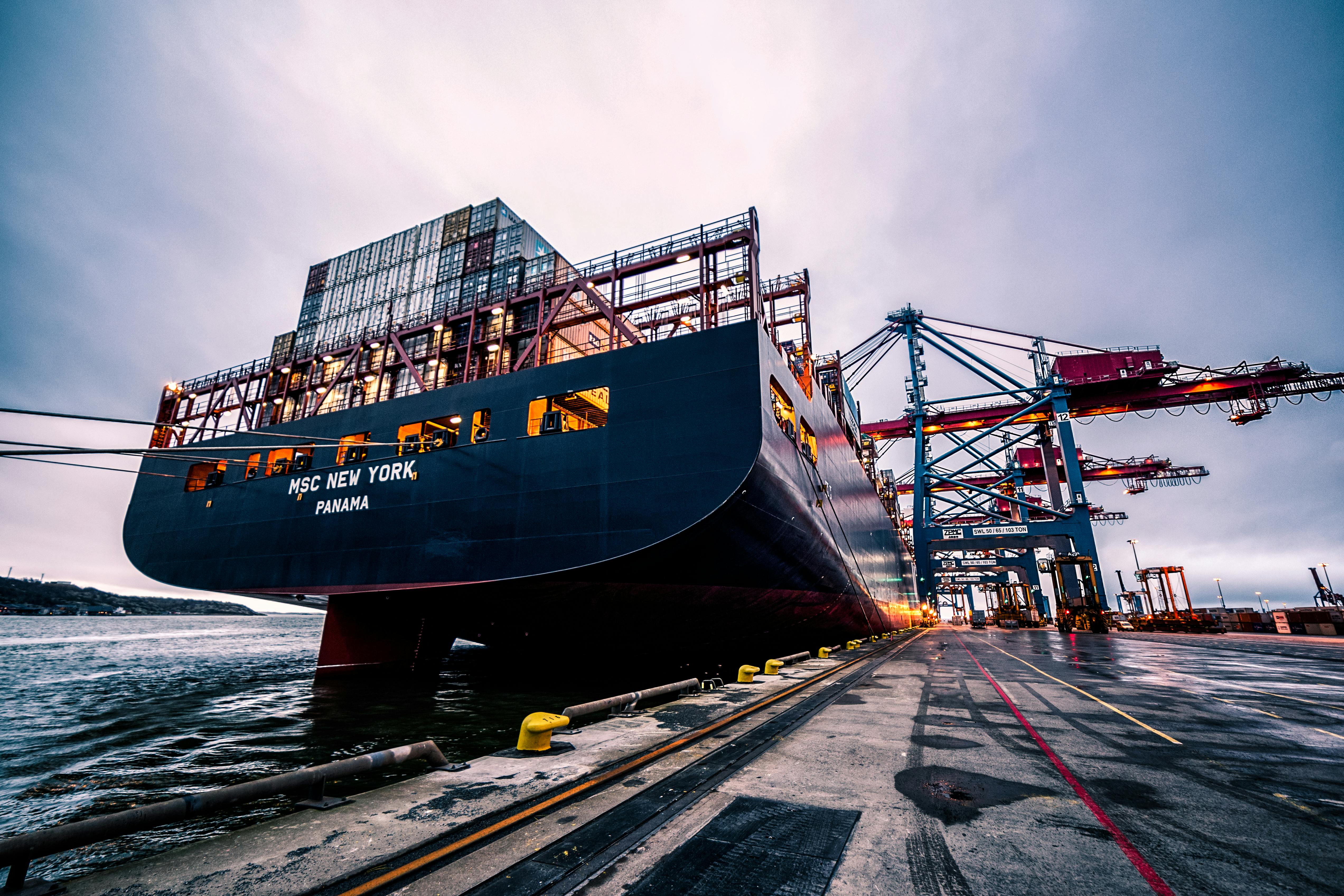 The ship is loaded with stacks of shipping containers and connected to the dock with thick ropes.