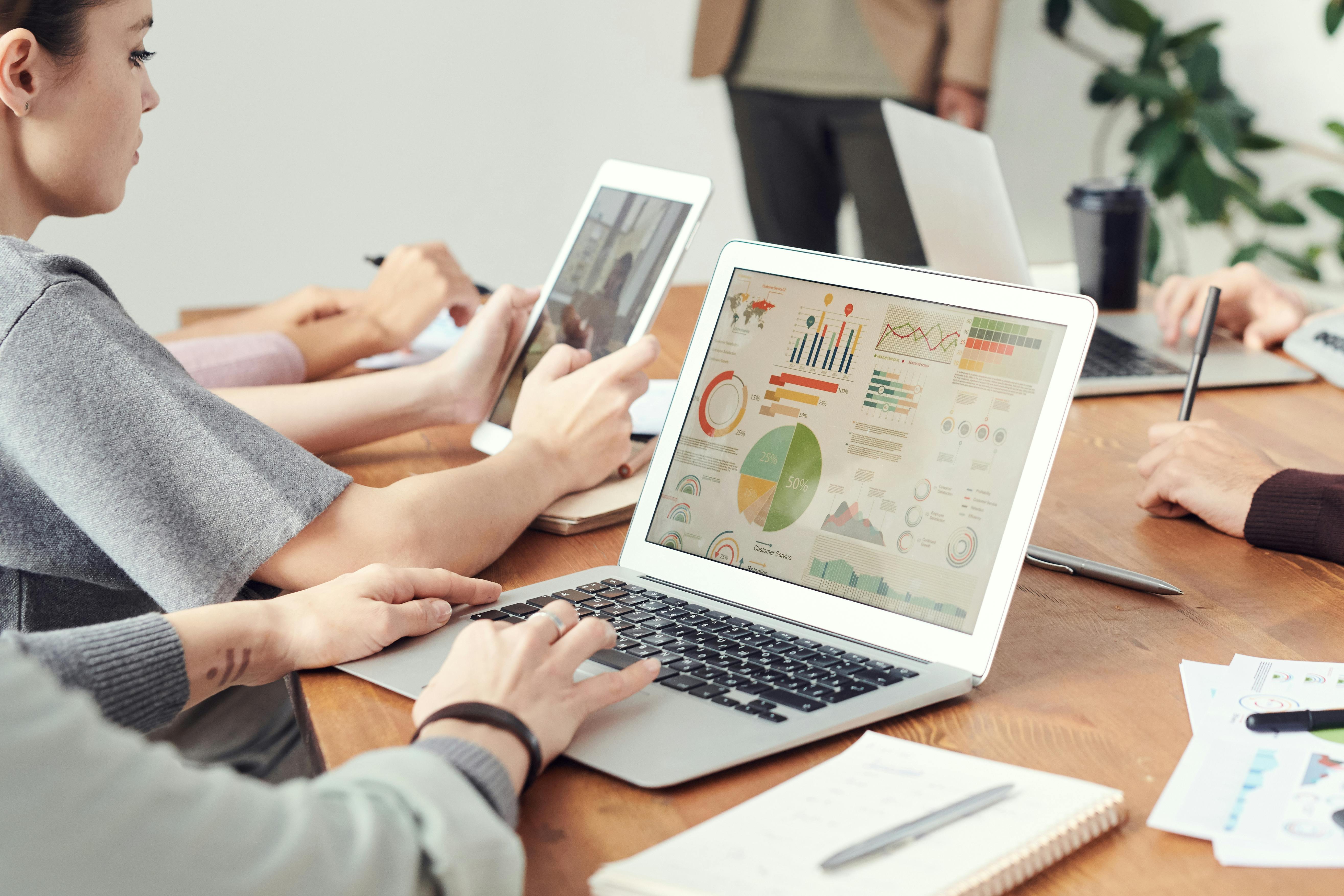 Group of people at a meeting table working on laptops and tablets displaying charts and graphs.