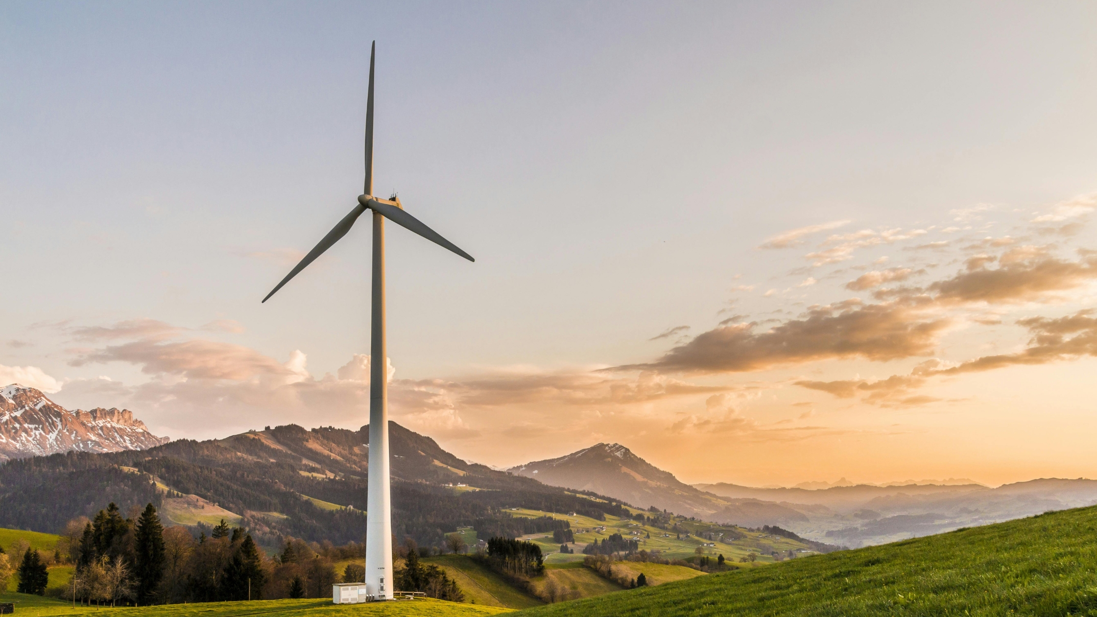 Wind turbine on a grassy hill with mountains at sunset.
