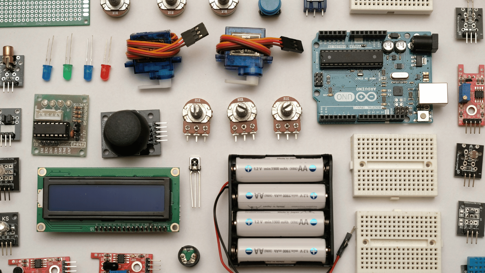A top-down, neatly organized arrangement of DIY electronic components on a white surface. The collection features an Arduino UNO microcontroller board, a green LCD screen, a four-AA battery pack, blue mini servo motors, white breadboards, potentiometers, colorful LEDs, and various small sensor modules.