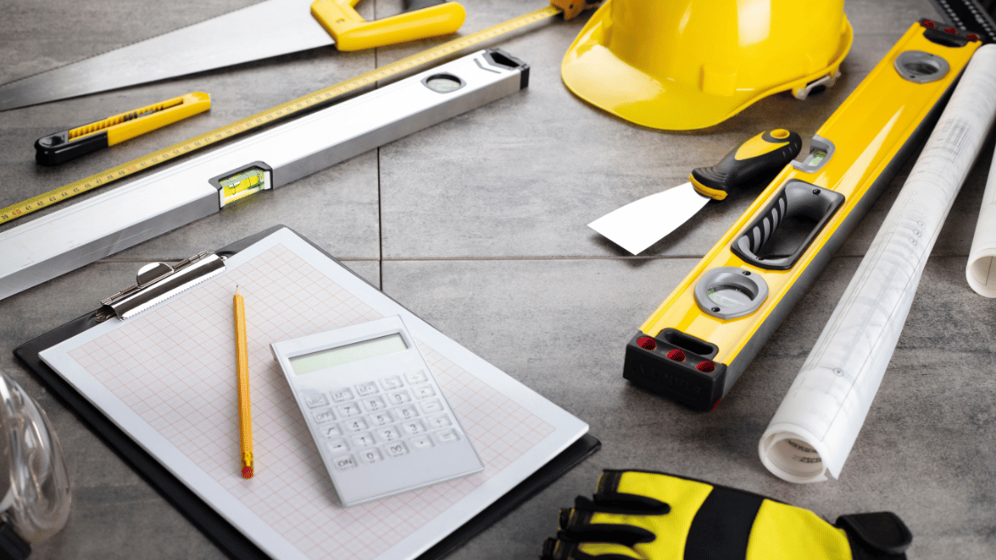 Assorted construction tools, blueprints, a hard hat, and a clipboard with a calculator arranged on a flat surface.