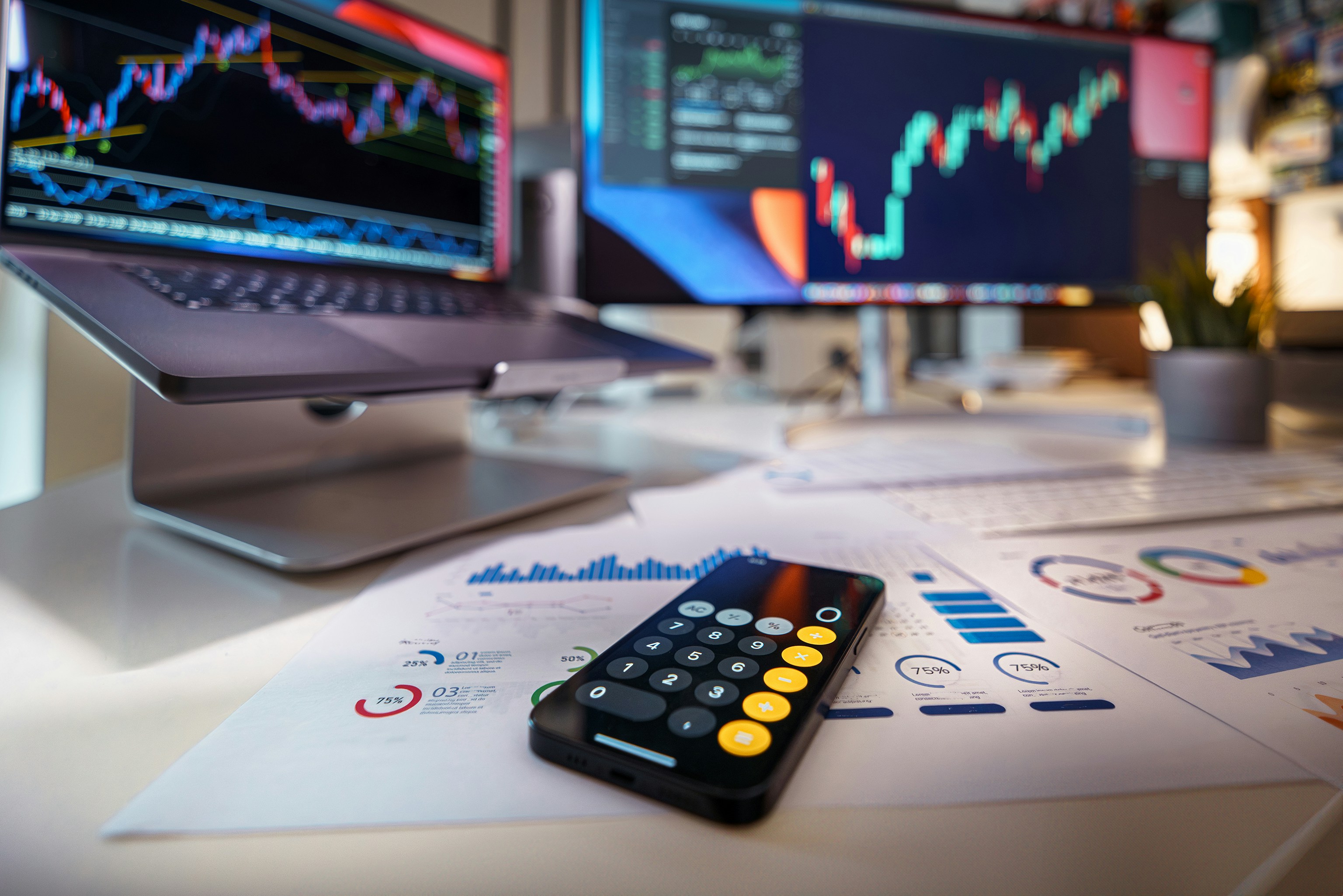 Close-up of a modern financial workspace featuring a smartphone with a calculator app open, financial charts and graphs on printed documents, and two computer screens displaying colorful stock market data and trend lines.