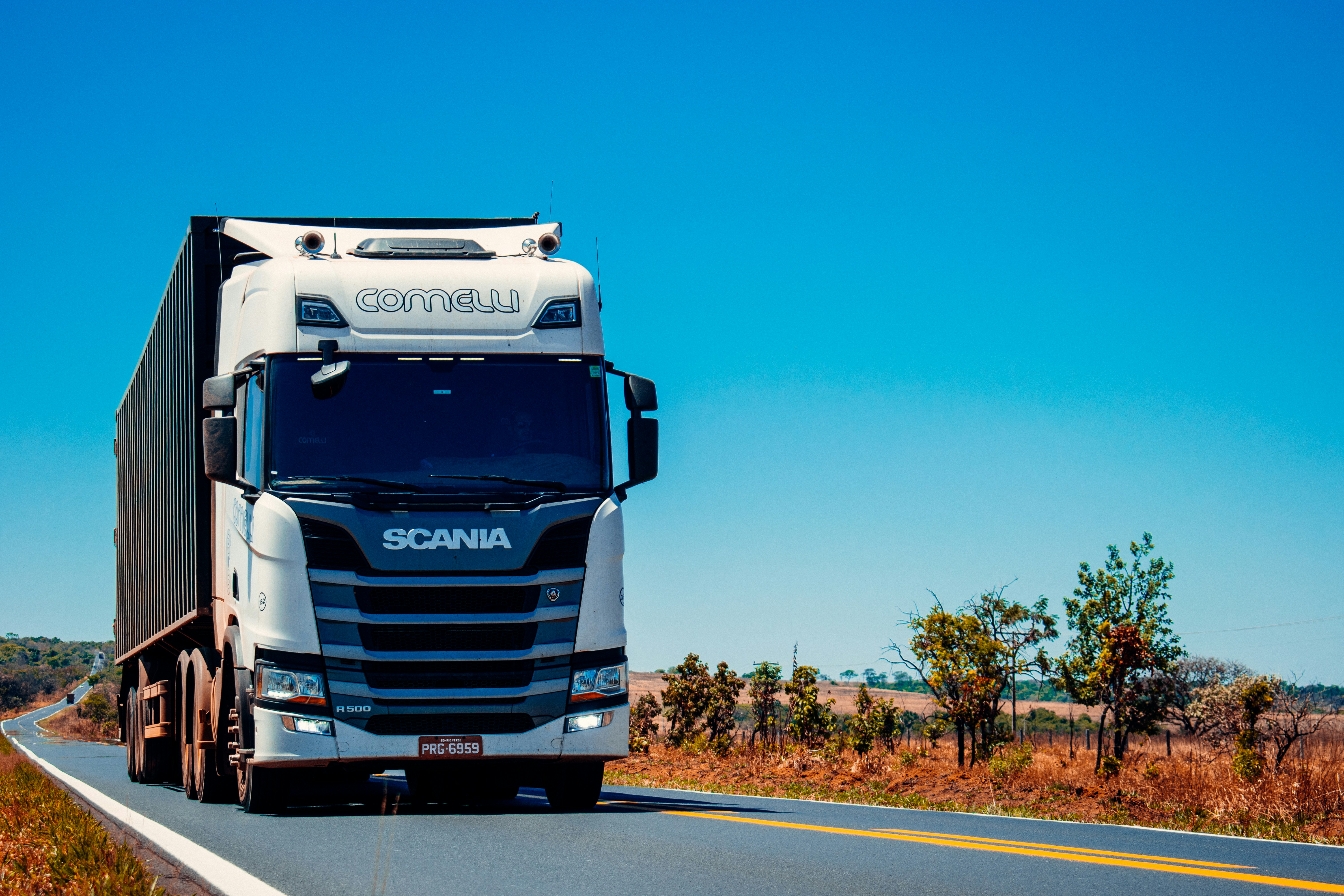White Scania semi-truck with a container trailer driving on a rural highway.