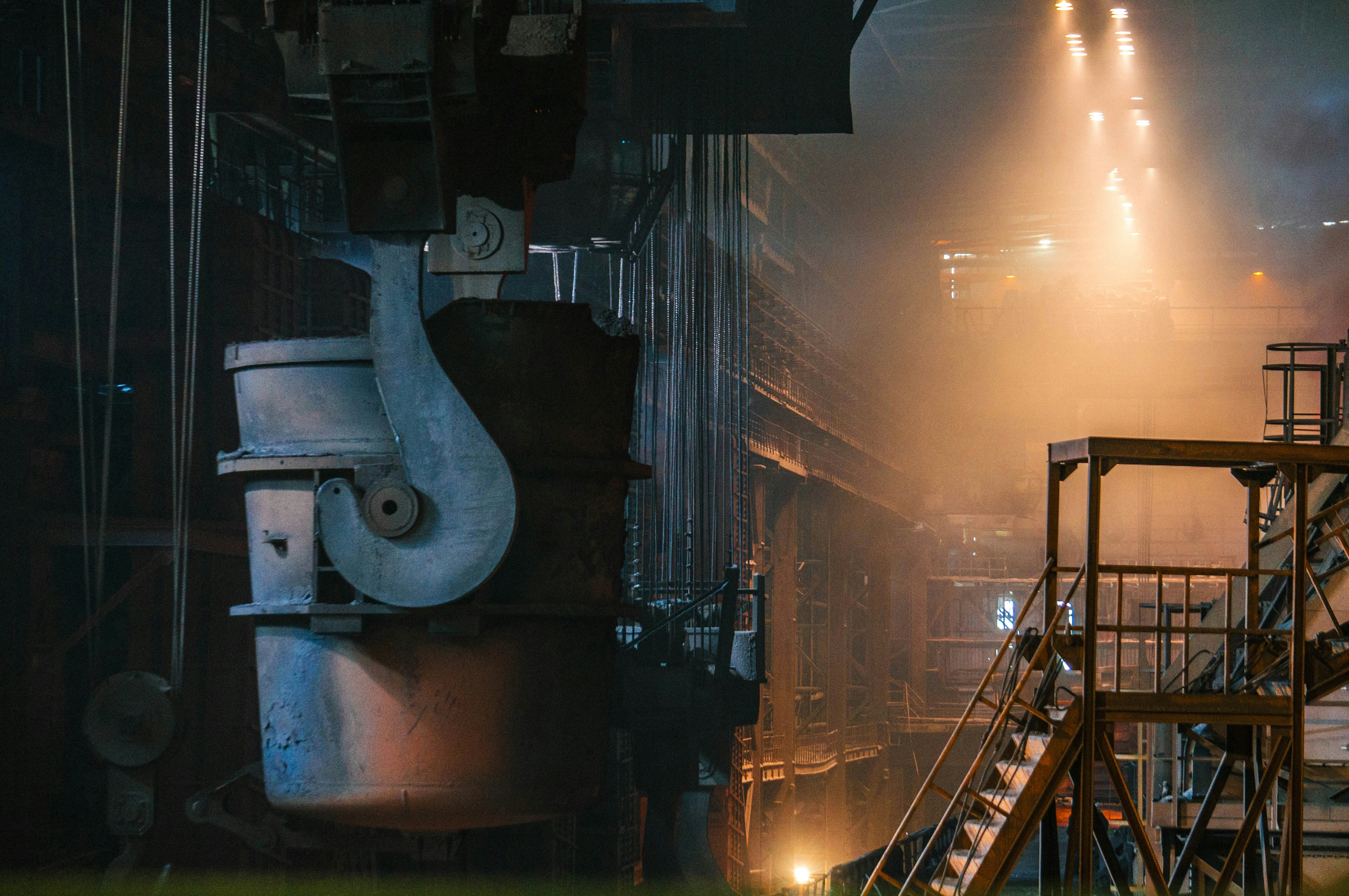 Large industrial steel foundry interior with a massive metal ladle suspended from a crane, surrounded by metal structures.