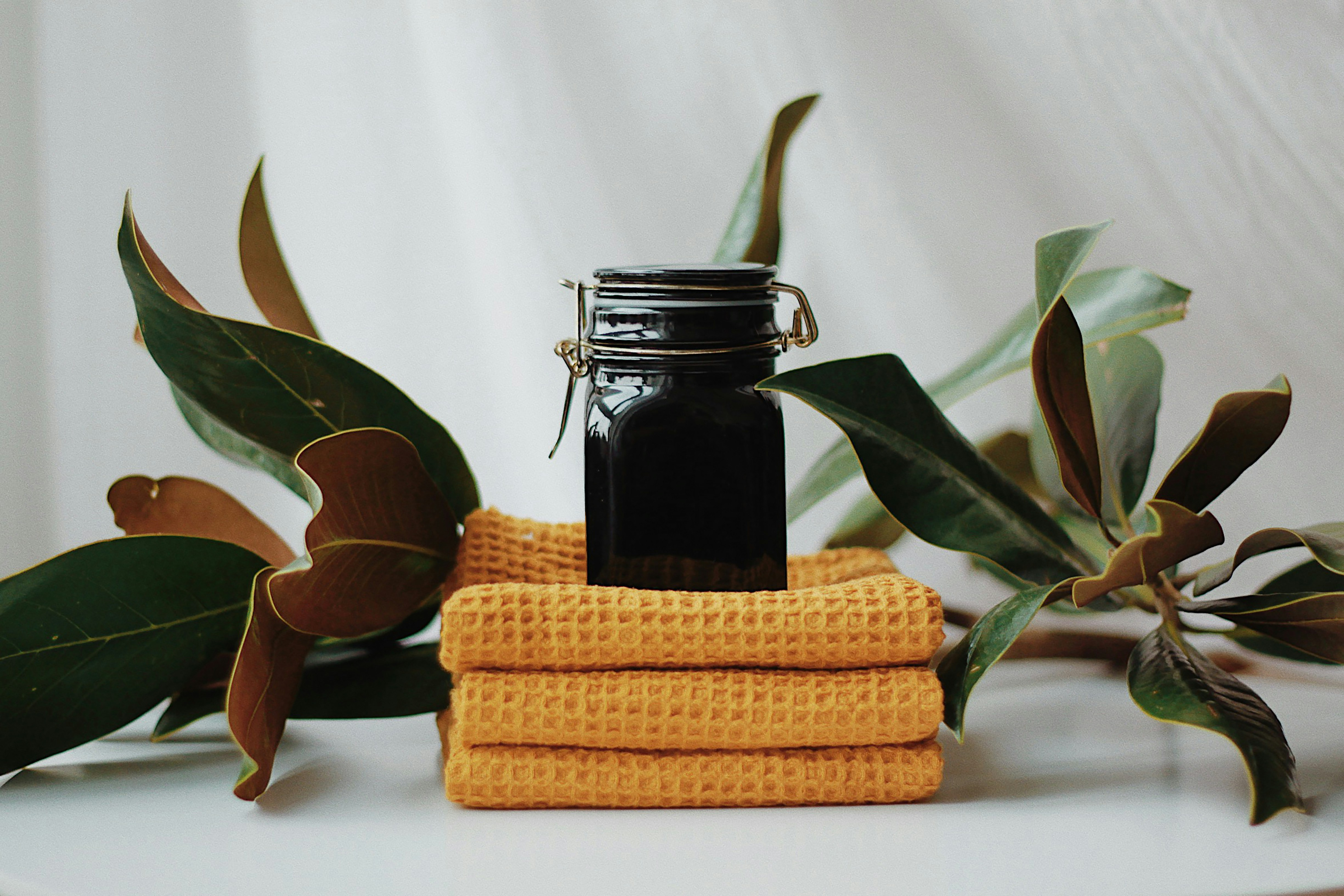 A small black glass jar sits atop three neatly folded yellow waffle-textured towels, surrounded by green leaves. The setting highlights themes of product purity and cosmetic safety.