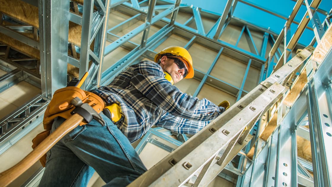 A construction worker wearing a yellow hard hat and tool belt climbs a metal ladder inside a building's steel framework.